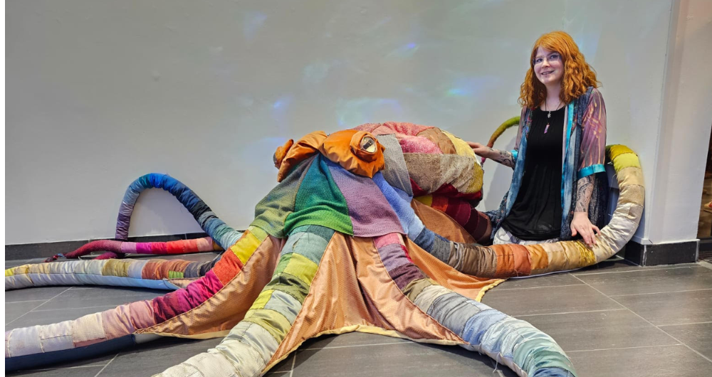 Artists in Residence Erin Redmond pictured sitting on the floor of the LAB Gallery next to a large fabric octopus stitched together from recycled swatches of fabric in rainbow colours