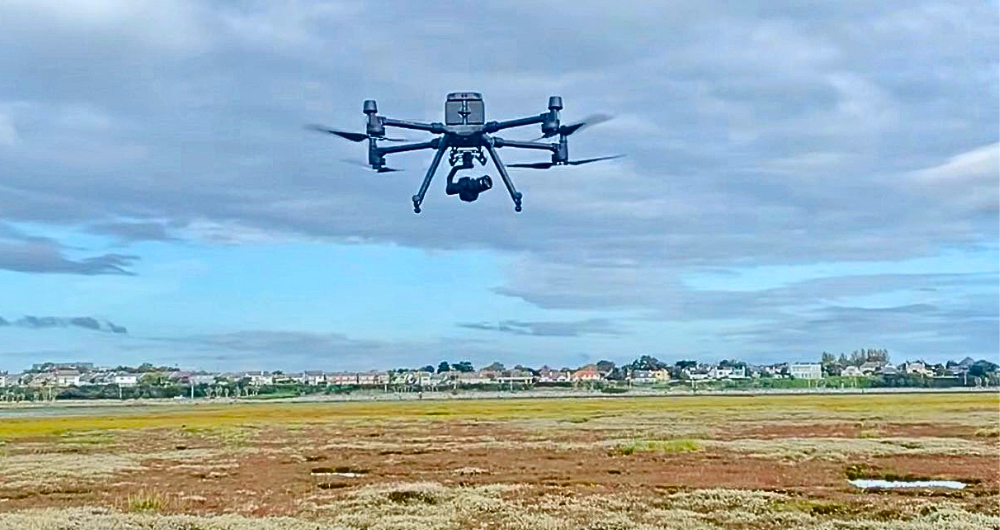 iHabimap drone hovering over scrubland with buildings on the horizon. Cloudy sky